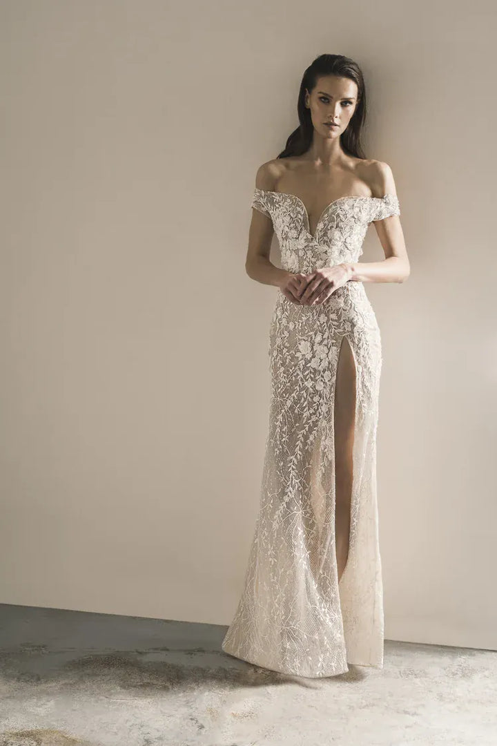 A woman modeling an off-the-shoulder, floor-length lace gown with a thigh-high slit, standing against a plain beige wall.
