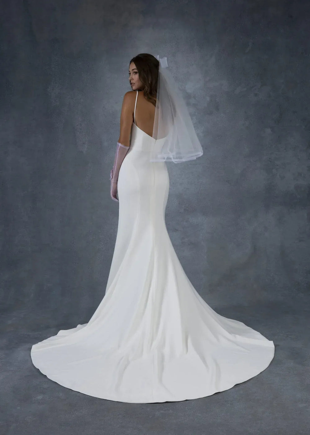 A woman modeling a white bridal gown from the back, wearing a sheer veil and long gloves, against a textured gray studio backdrop.