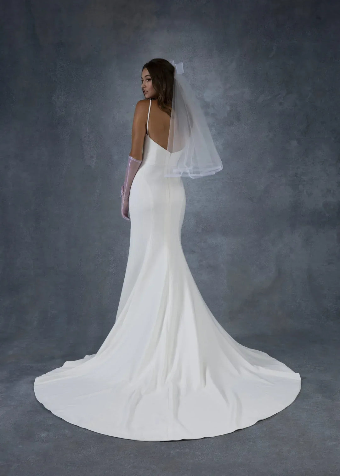 A woman modeling a white bridal gown from the back, wearing a sheer veil and long gloves, against a textured gray studio backdrop.