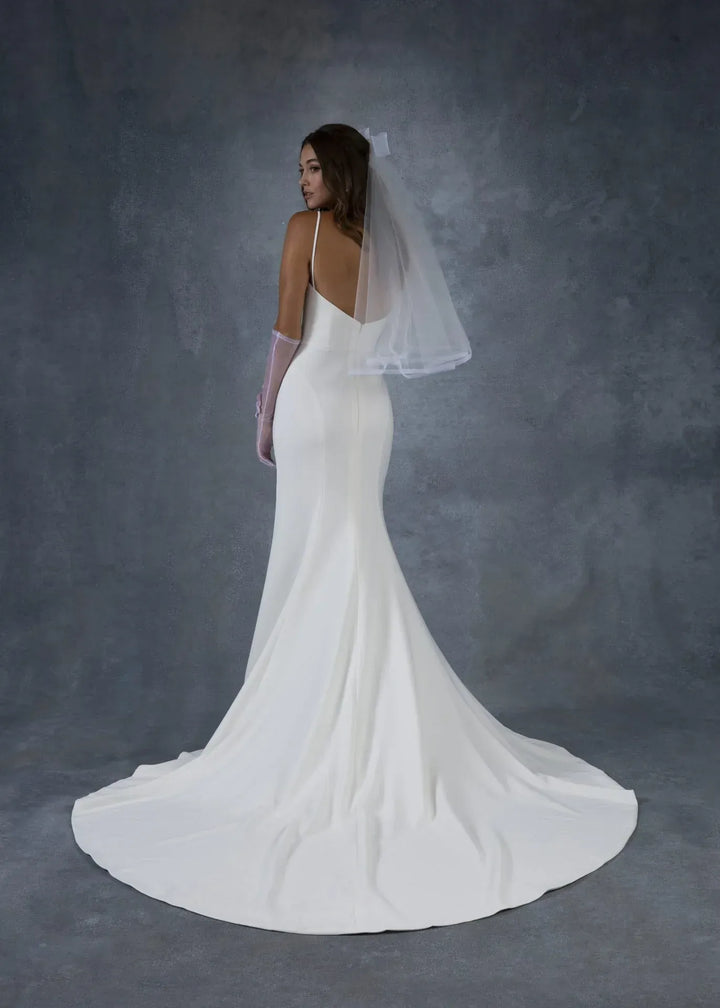 A woman modeling a white bridal gown from the back, wearing a sheer veil and long gloves, against a textured gray studio backdrop.