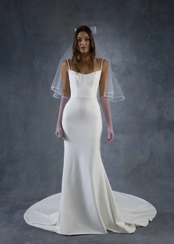 A woman modeling a sleeveless white wedding gown with a fitted bodice, flared skirt, and long sheer veil against a dark gray studio backdrop.