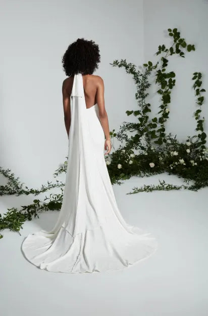 A woman stands with her back to the camera wearing a sleeveless white bridal gown with a long draped train and open back. The setting is a bright white studio with green vine decor along the wall and floor.