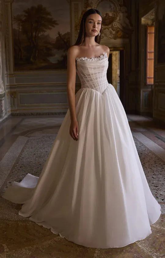 A woman wearing a strapless white ball gown with a structured corset-style bodice and full flowing skirt, standing in an ornate interior room with classical wall art and stone flooring.
