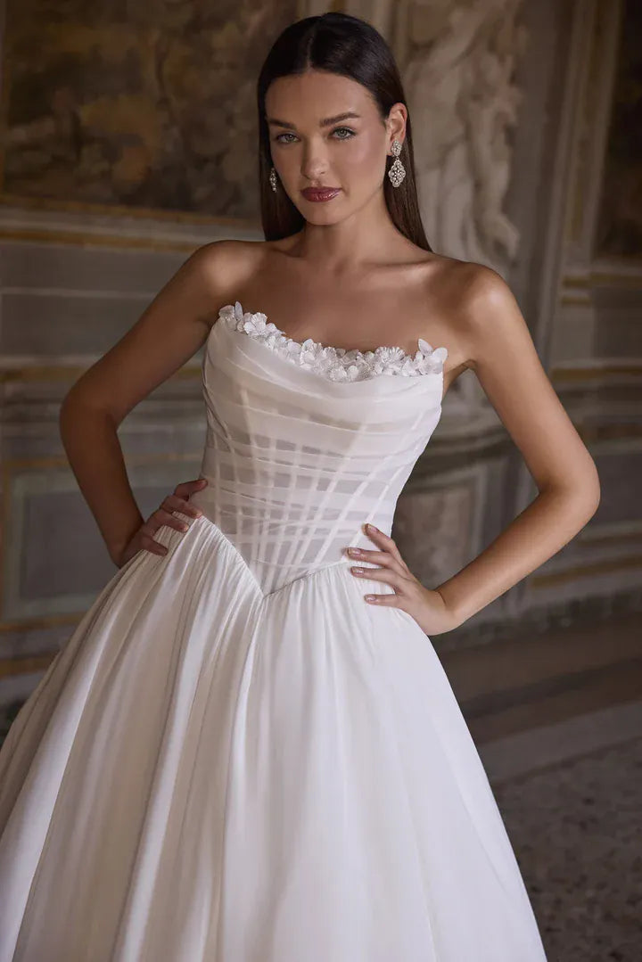 A woman modeling a strapless white wedding gown with a structured corset-style bodice and full skirt, posed indoors against a classical stone wall.