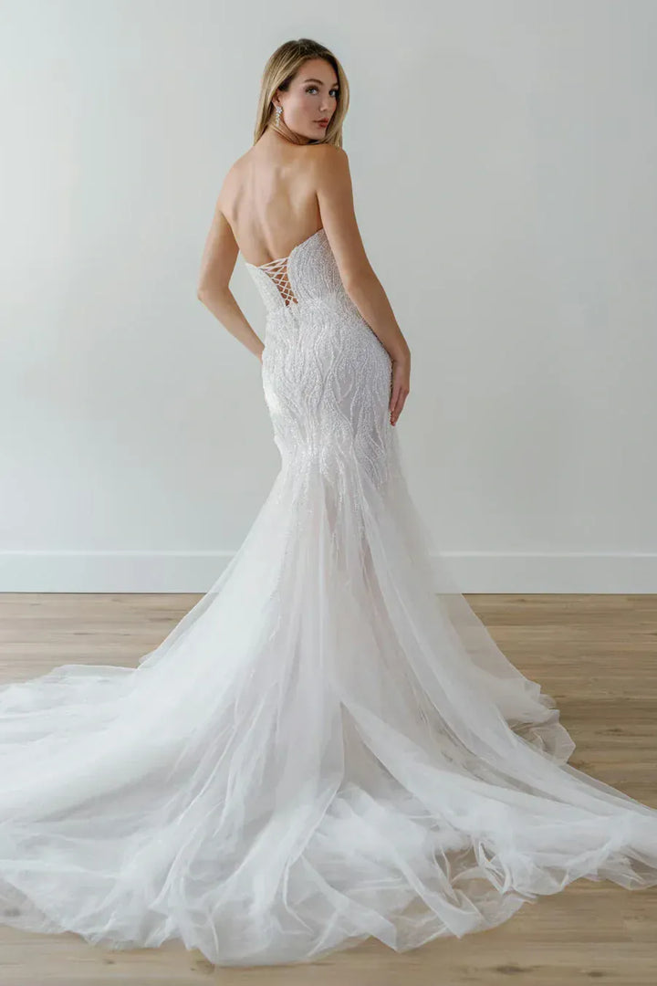 A woman modeling a strapless white lace mermaid wedding gown with a lace-up open back and a long tulle train, photographed in a bright indoor studio with a light wall and wood floor.