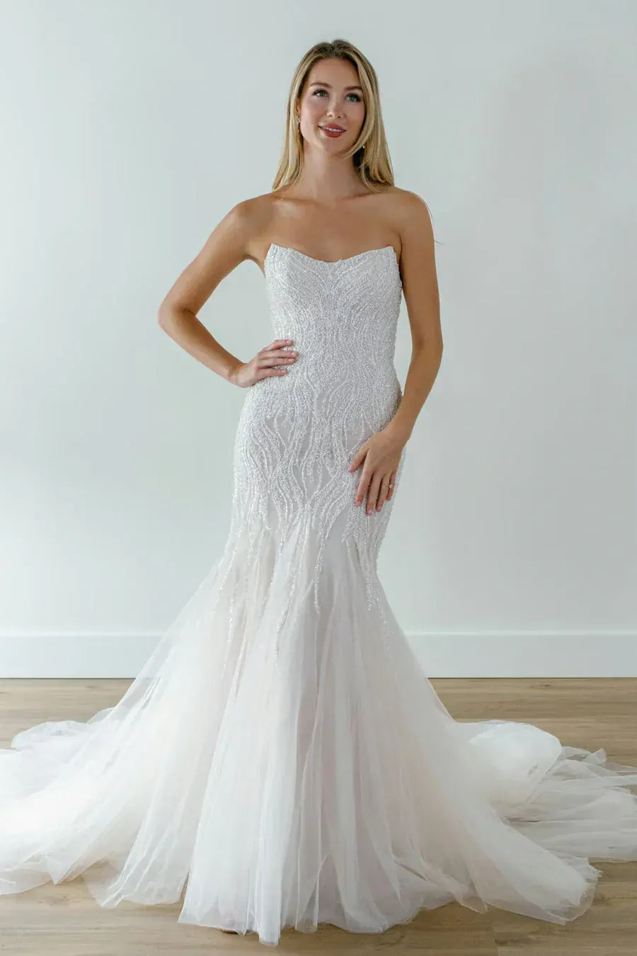 A woman modeling a strapless white mermaid-style wedding gown with intricate lace applique on the bodice and a layered tulle skirt with a train, standing against a plain light wall and wood floor.