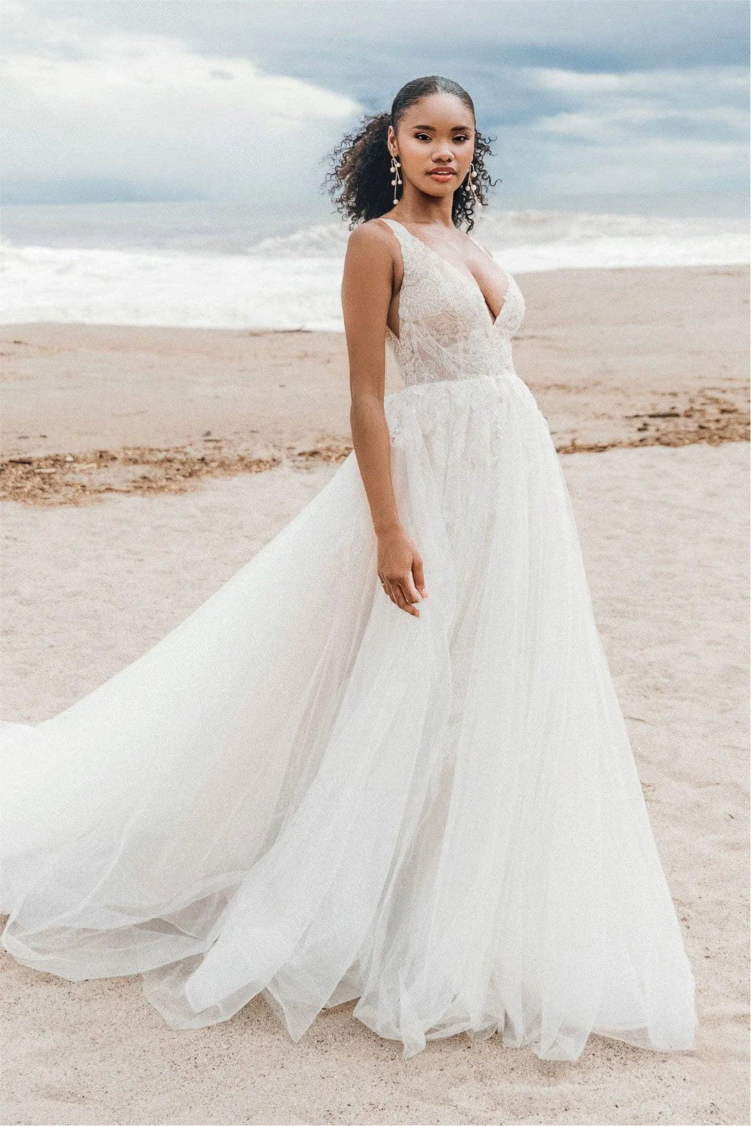 Woman in a white wedding dress standing on a beach with a cloudy sky.