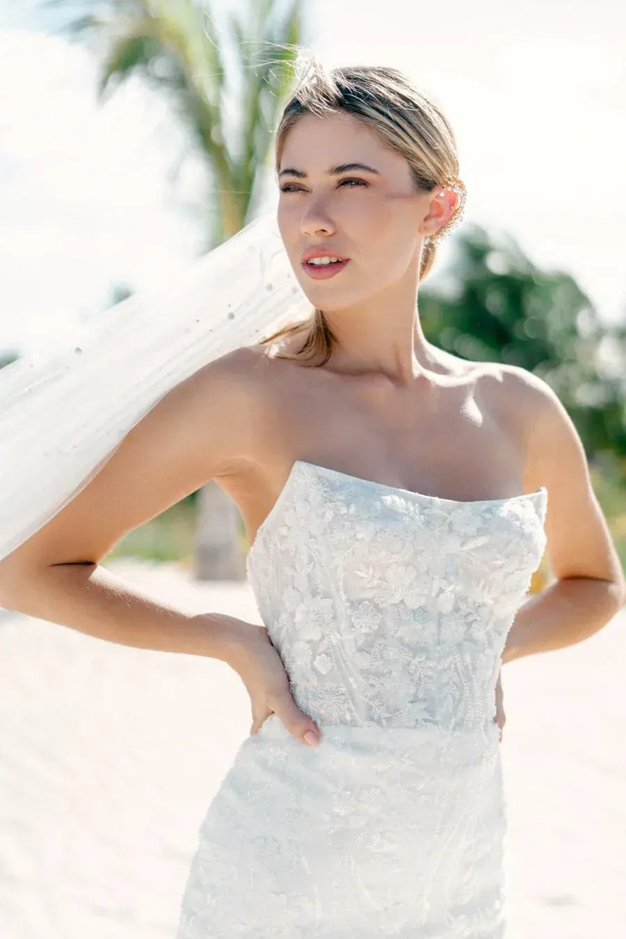 A woman wearing a strapless white lace wedding gown and veil, standing outdoors on a bright sandy beach with blurred palm trees in the background.