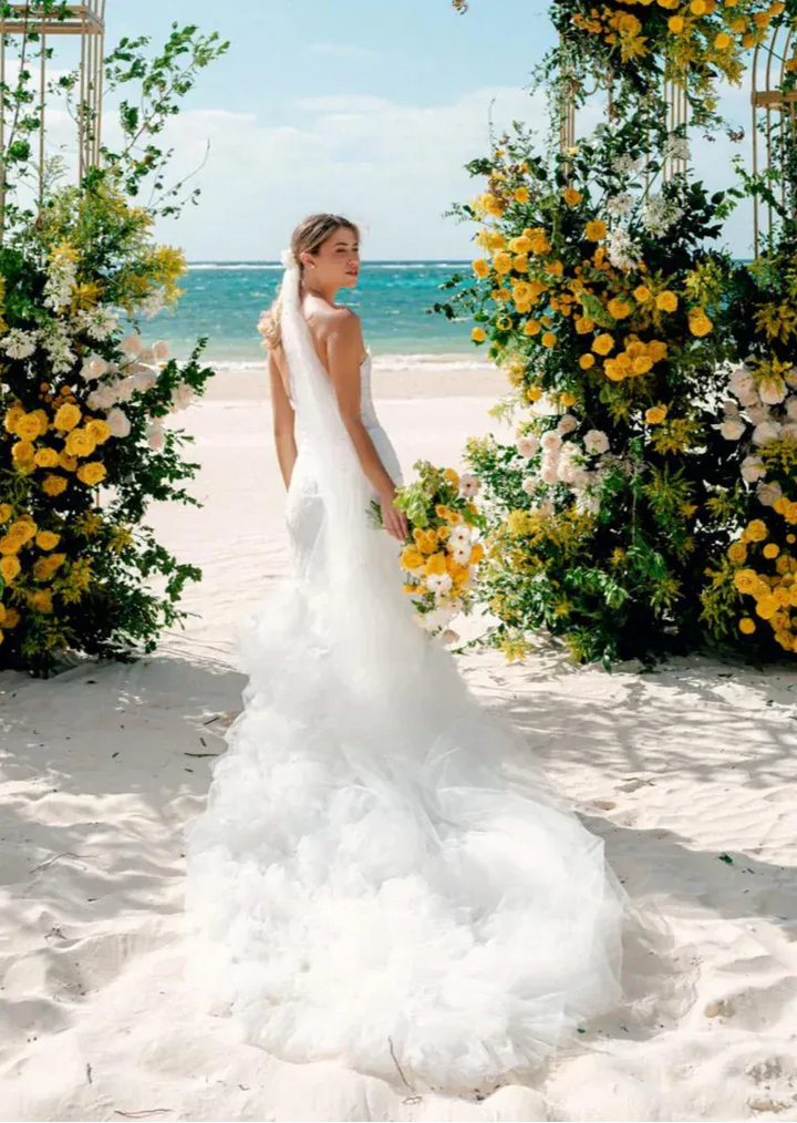 A woman in a white wedding gown stands on a sandy beach between large floral arches with yellow and white flowers, with the ocean and blue sky in the background.