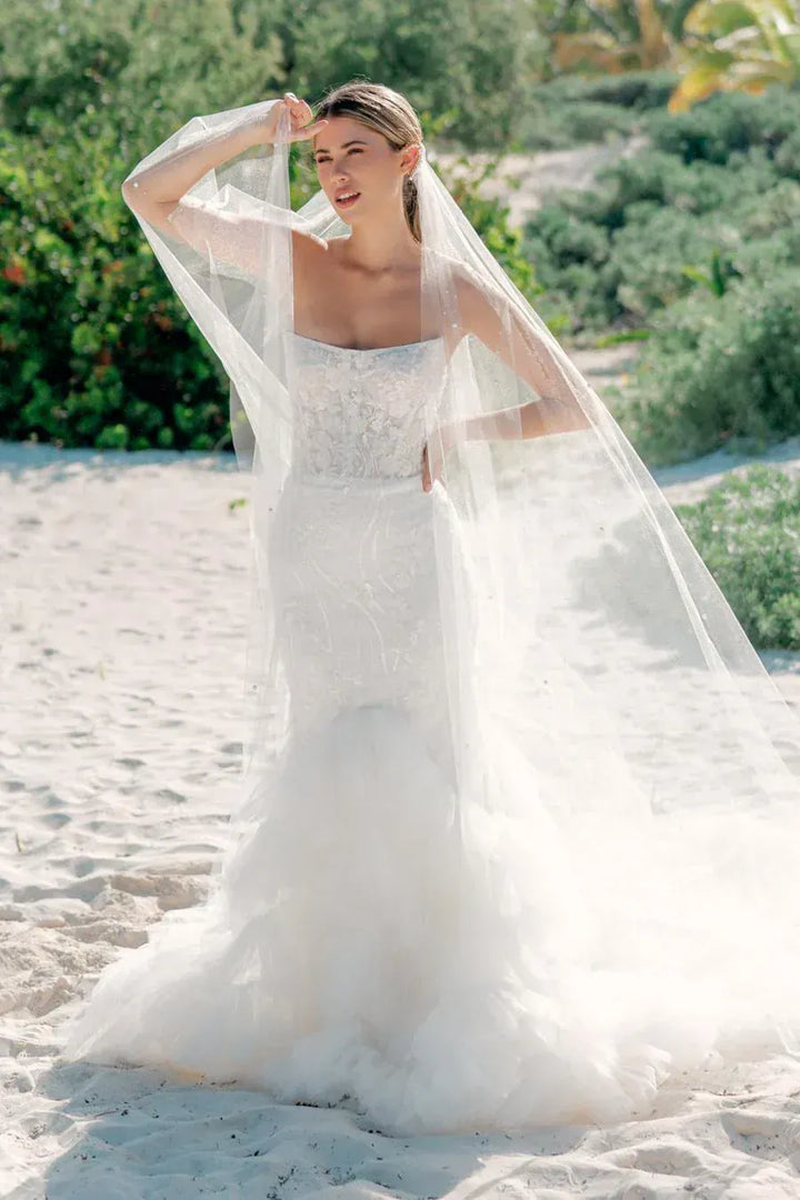 A woman wearing a white strapless lace wedding gown with a long sheer veil stands on a sandy beach with green foliage in the background.