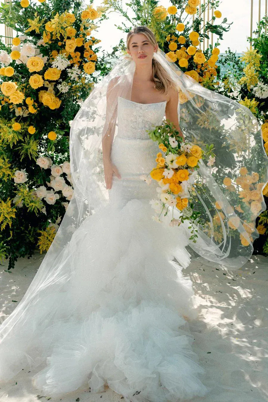 A bride in a strapless white tulle wedding gown with a long sheer veil and a bouquet of yellow and white flowers, standing in front of a large floral backdrop of yellow roses and greenery.