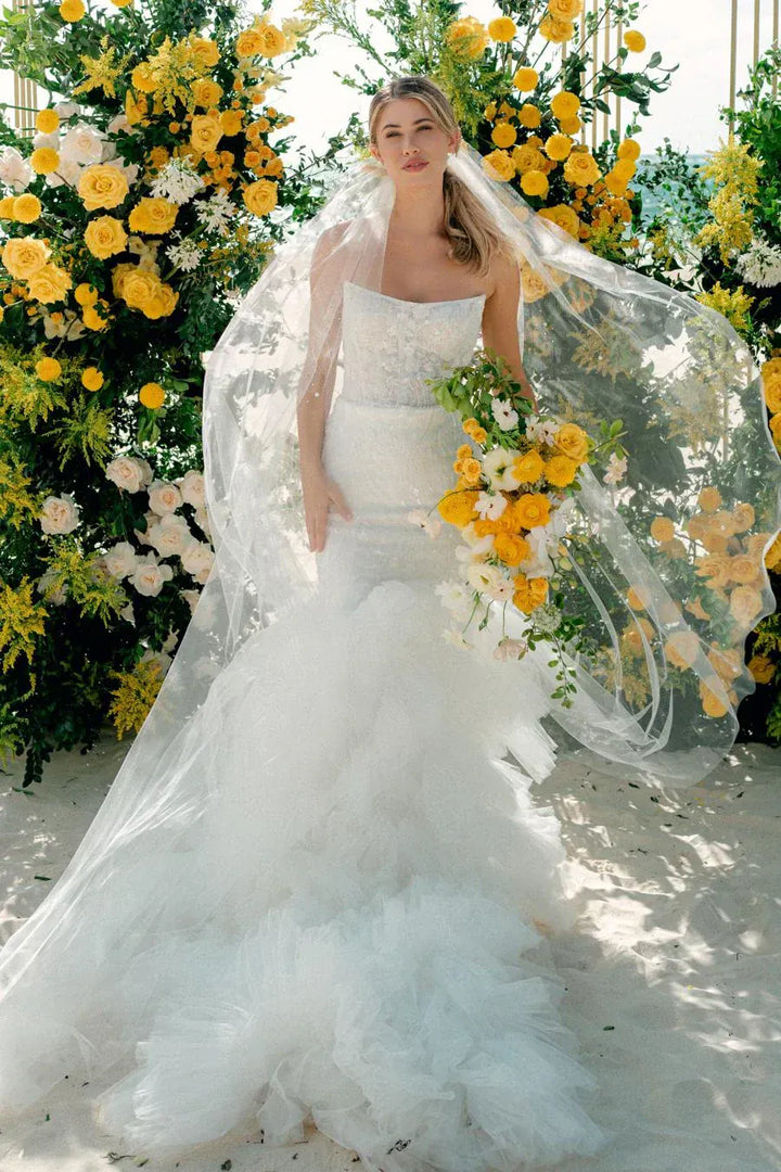 A bride in a strapless white tulle wedding gown with a long sheer veil and a bouquet of yellow and white flowers, standing in front of a large floral backdrop of yellow roses and greenery.