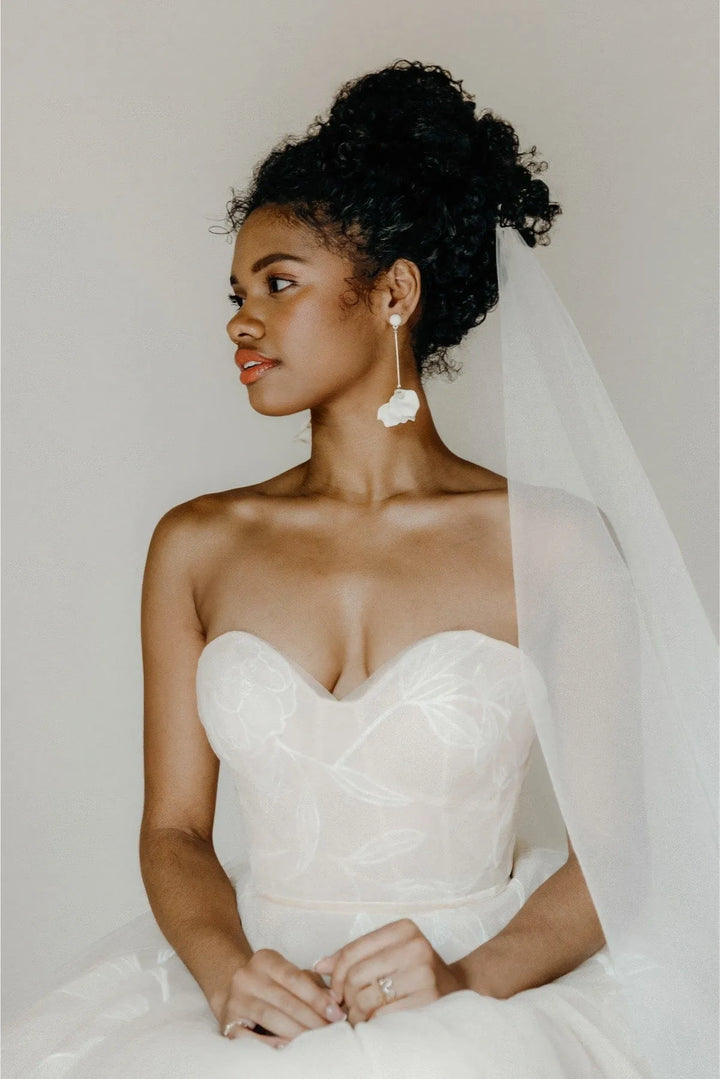 A woman in a strapless white wedding gown with a sheer veil and long white flower drop earrings, posed against a soft light gray studio background.