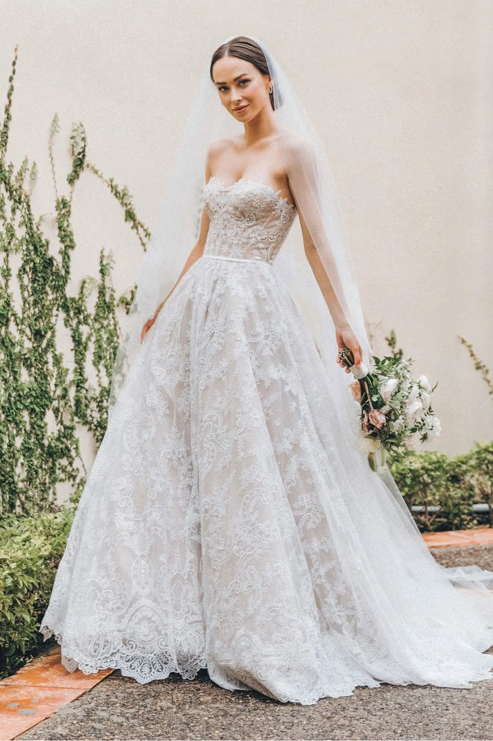 Woman in a white lace wedding dress standing outdoors with greenery in the background