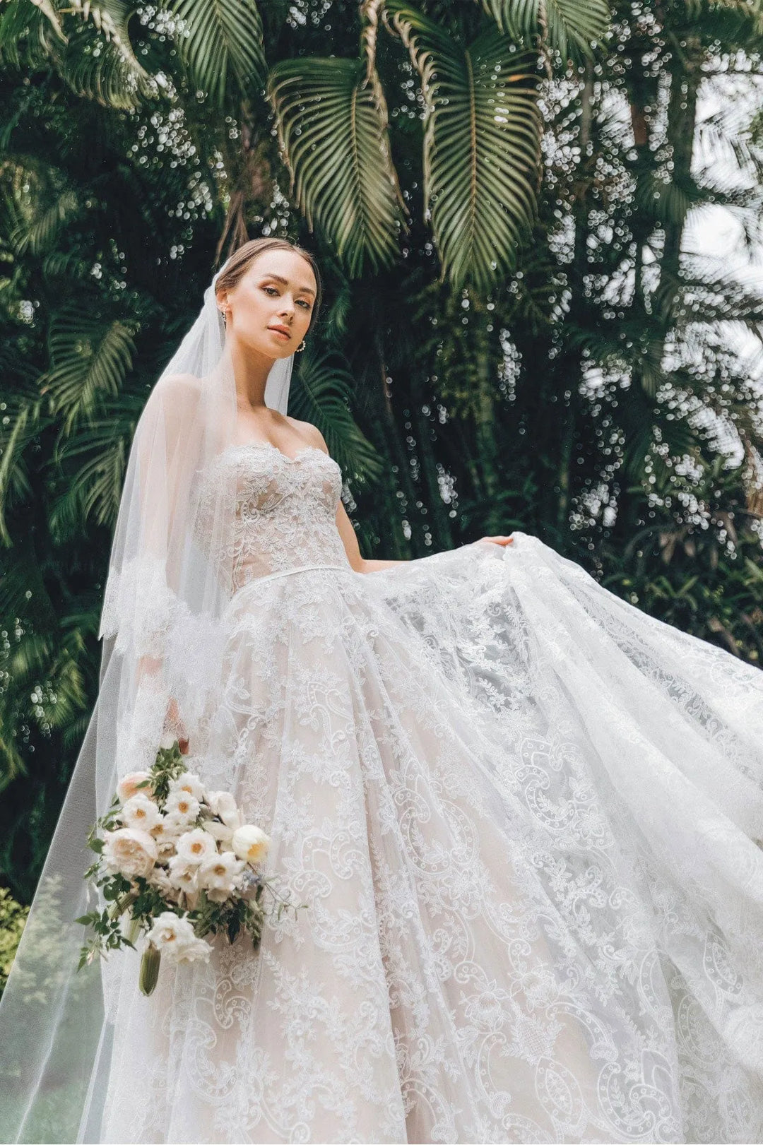 Bride in a white wedding dress with a long veil and bouquet standing in front of green foliage.