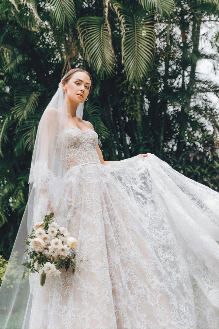 Bride in a white wedding dress with a long veil and bouquet standing in front of green foliage.