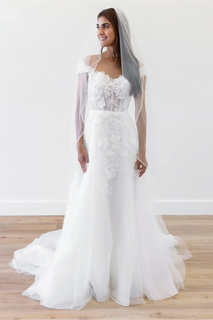 Woman wearing a white lace wedding dress with a sheer overlay on a plain background