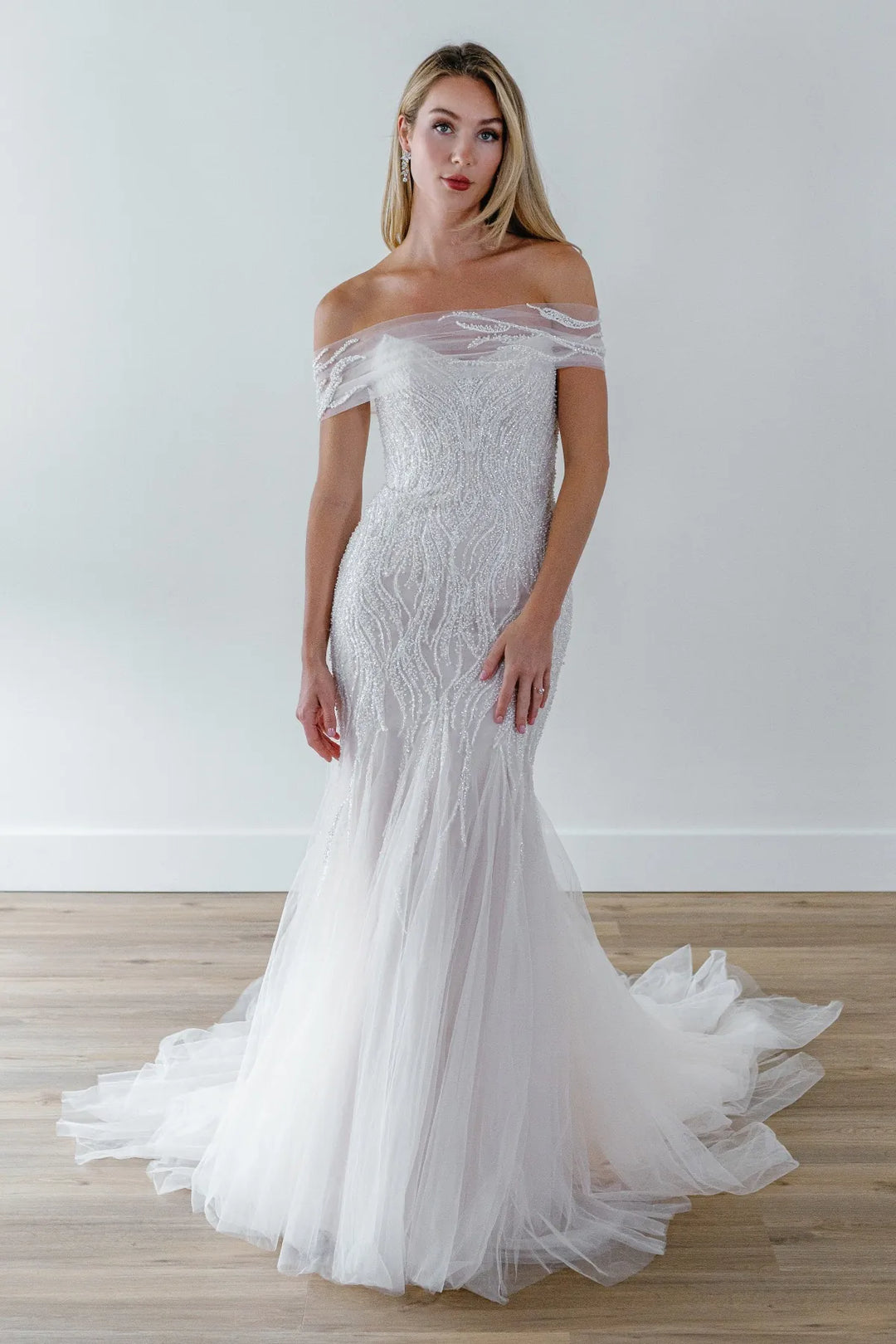 A woman modeling an off-the-shoulder white mermaid-style wedding gown with a fitted lace bodice and a full tulle mermaid skirt with a train, standing on a light wood floor against a plain light gray wall.