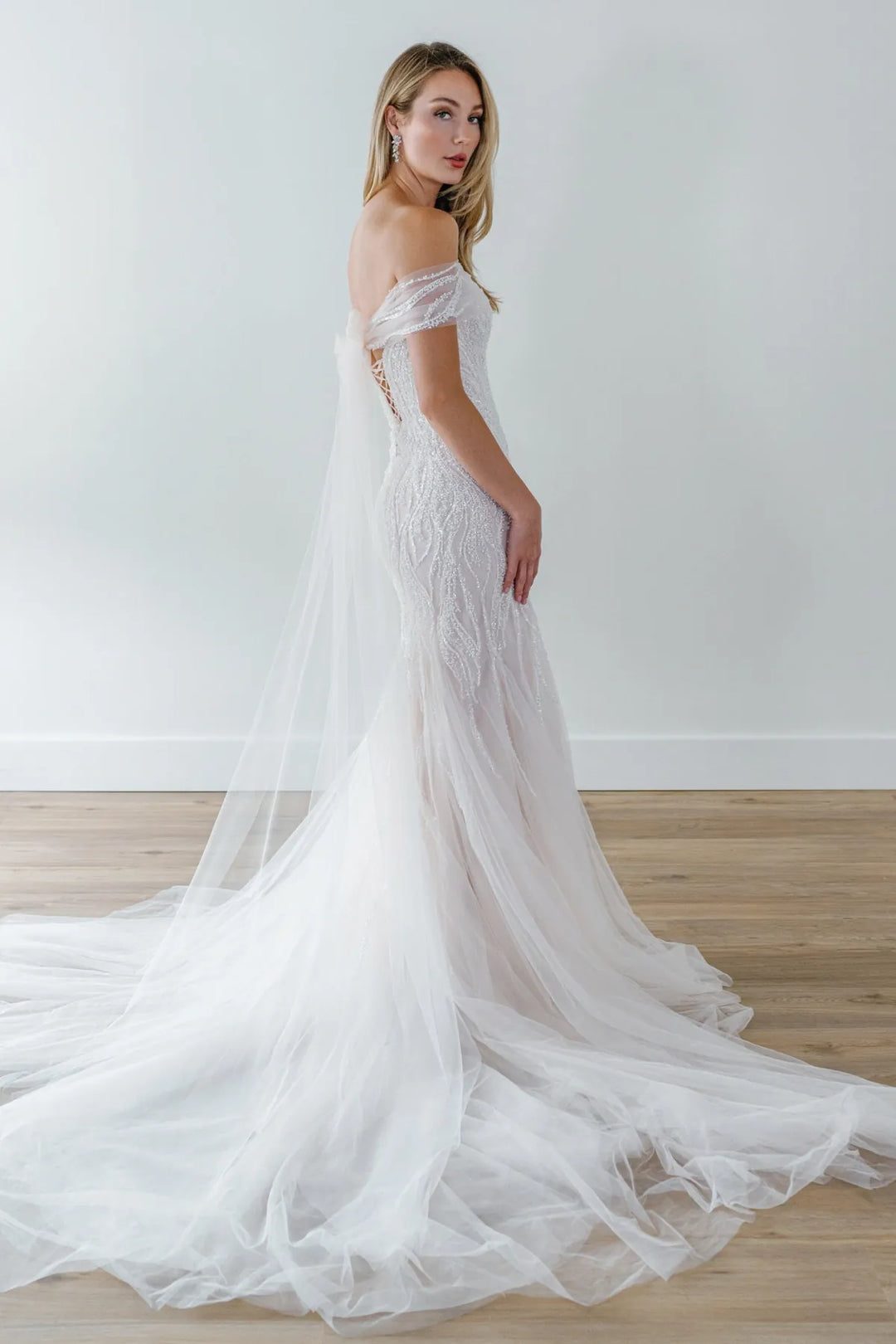 A woman modeling an off-the-shoulder white wedding gown with a long sheer tulle train and a matching veil, standing in a bright room with a light wall and wood floor.