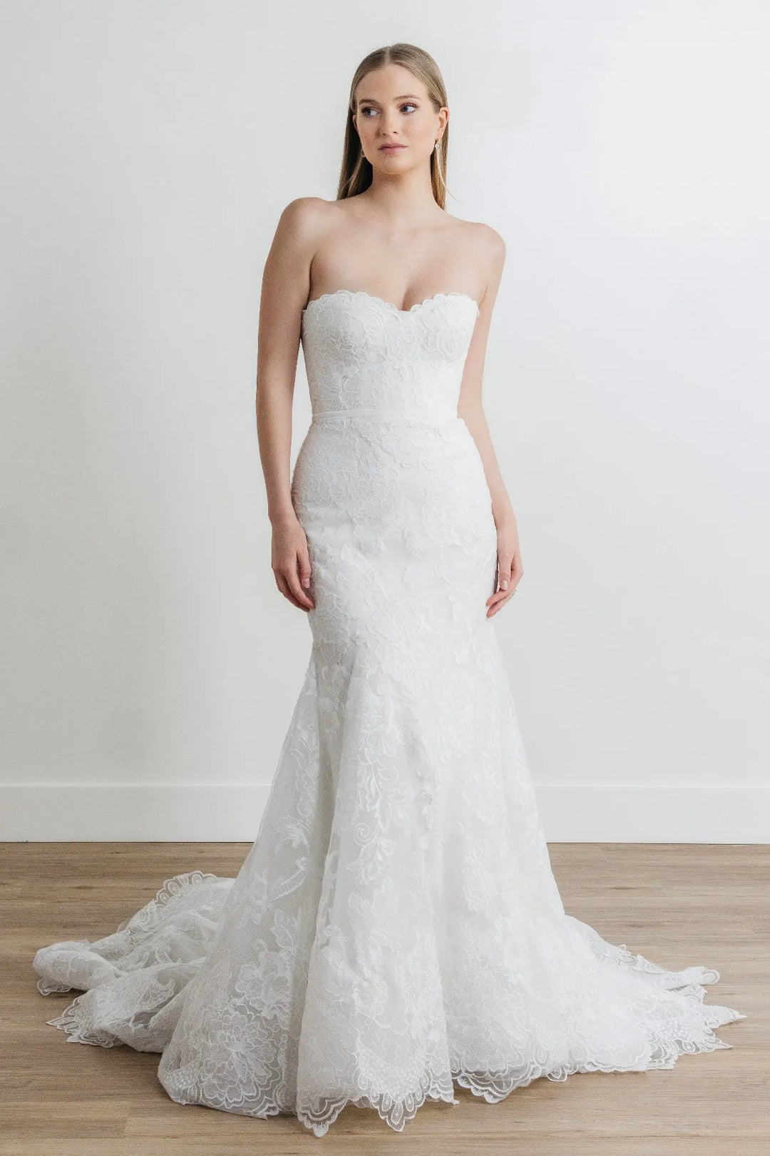 A woman modeling a strapless white lace mermaid-style wedding gown with a sweetheart neckline, fitted bodice, flared lace skirt, and a short train, standing against a plain light wall on a wood floor.