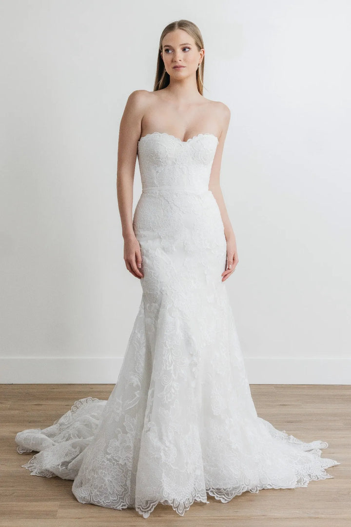A woman modeling a strapless white lace mermaid-style wedding gown with a sweetheart neckline, fitted bodice, flared lace skirt, and a short train, standing against a plain light wall on a wood floor.