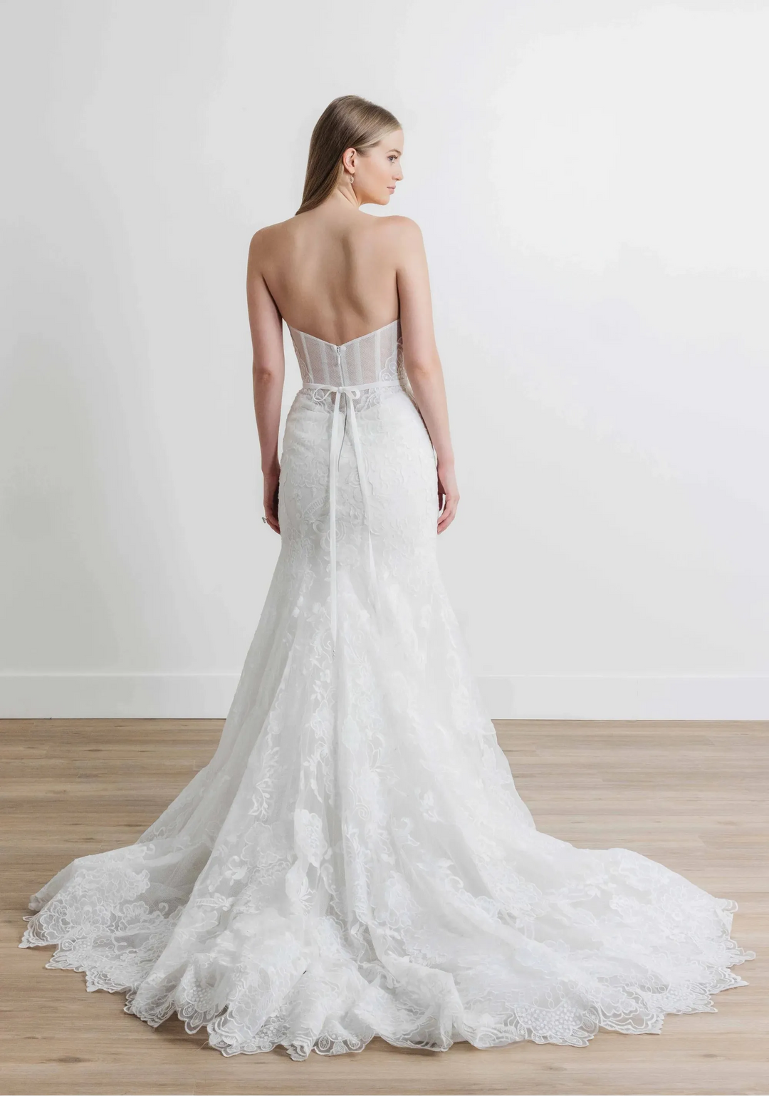 A woman modeling a strapless white lace wedding gown from the back, standing on a light wood floor against a plain white wall.