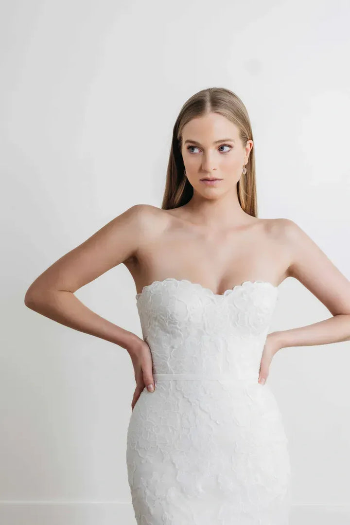 A woman modeling a strapless white lace wedding dress against a plain light gray studio background.