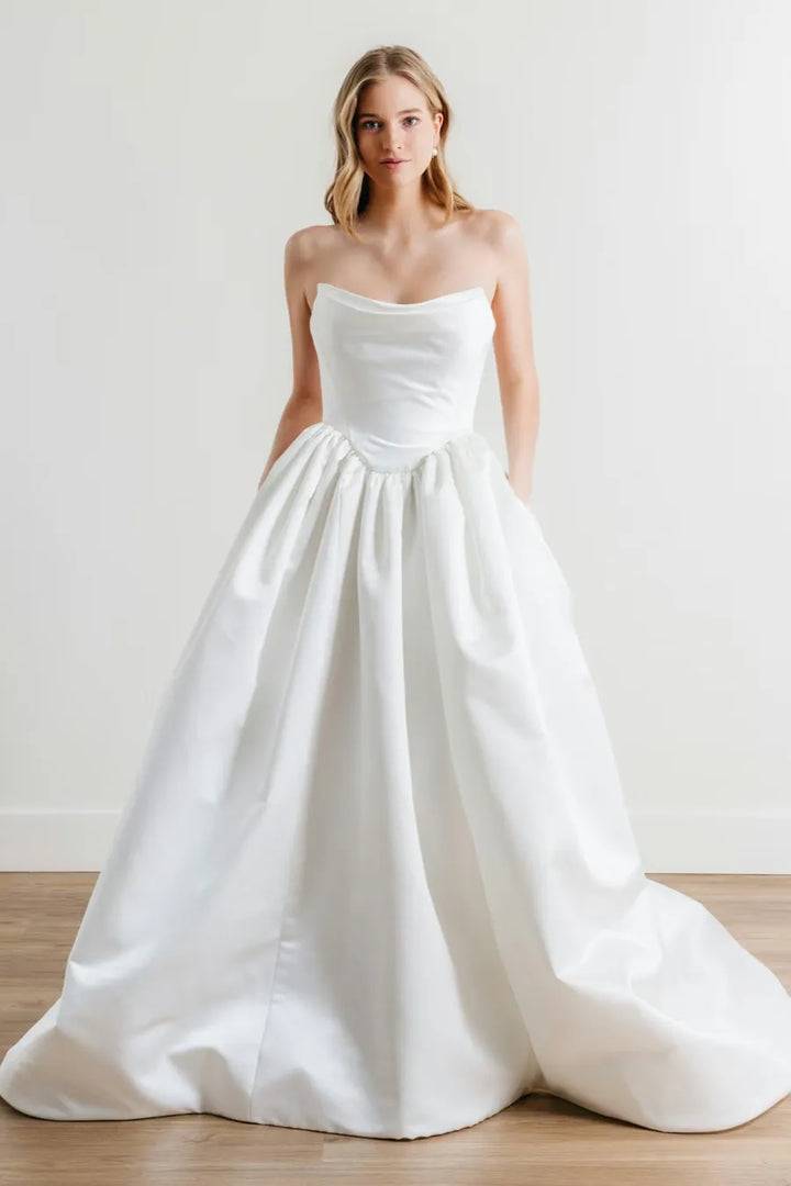 A woman modeling a strapless white ball gown wedding dress with a structured bodice and full skirt, standing on a light wood floor against a plain off-white wall.