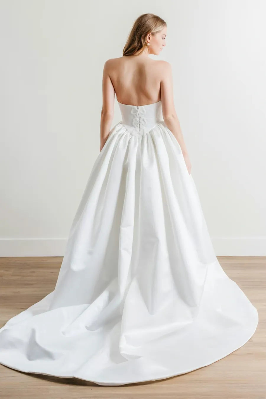 A woman wearing a strapless white wedding gown with a structured corset-style waist and a full satin train, photographed from the back in a bright indoor studio with a light wall and wood floor.