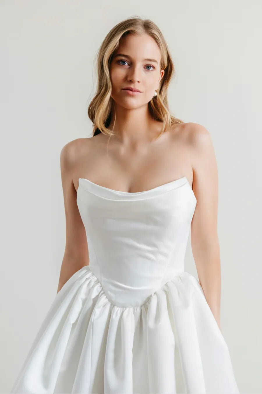 A woman wearing a strapless white dress with a structured sweetheart neckline and a full gathered skirt, photographed against a plain light gray studio background.