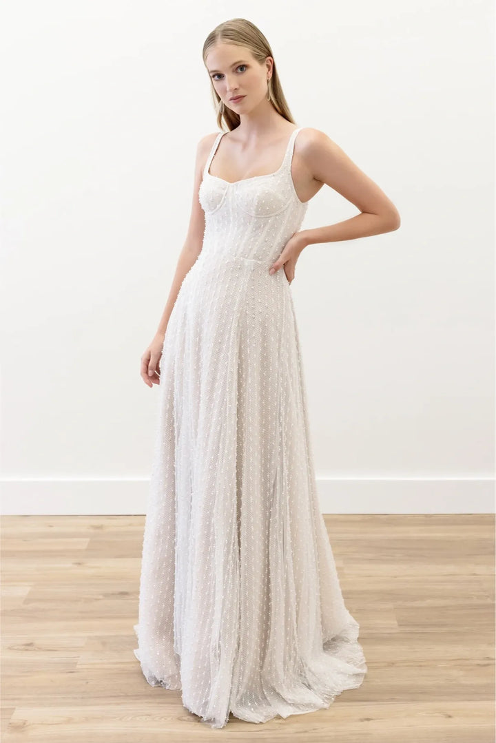 A woman modeling a sleeveless, floor-length white textured gown with a fitted bodice and flowing skirt, standing in a bright indoor room with a white wall and light wood floor.
