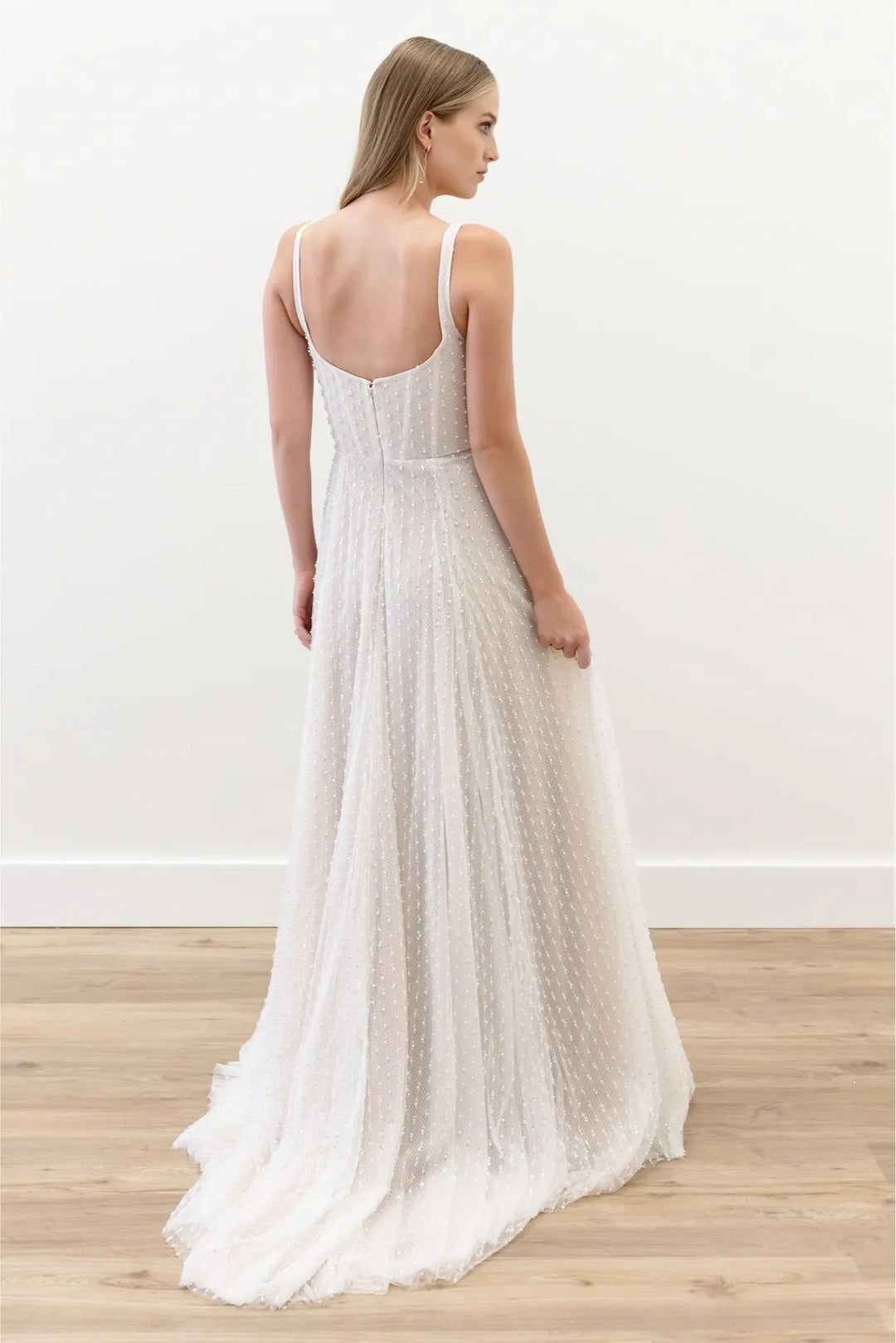 A woman wearing a sleeveless white maxi dress with thin straps, a fitted bodice, and a flowing textured skirt, shown from the back in a bright indoor room with a light wood floor.