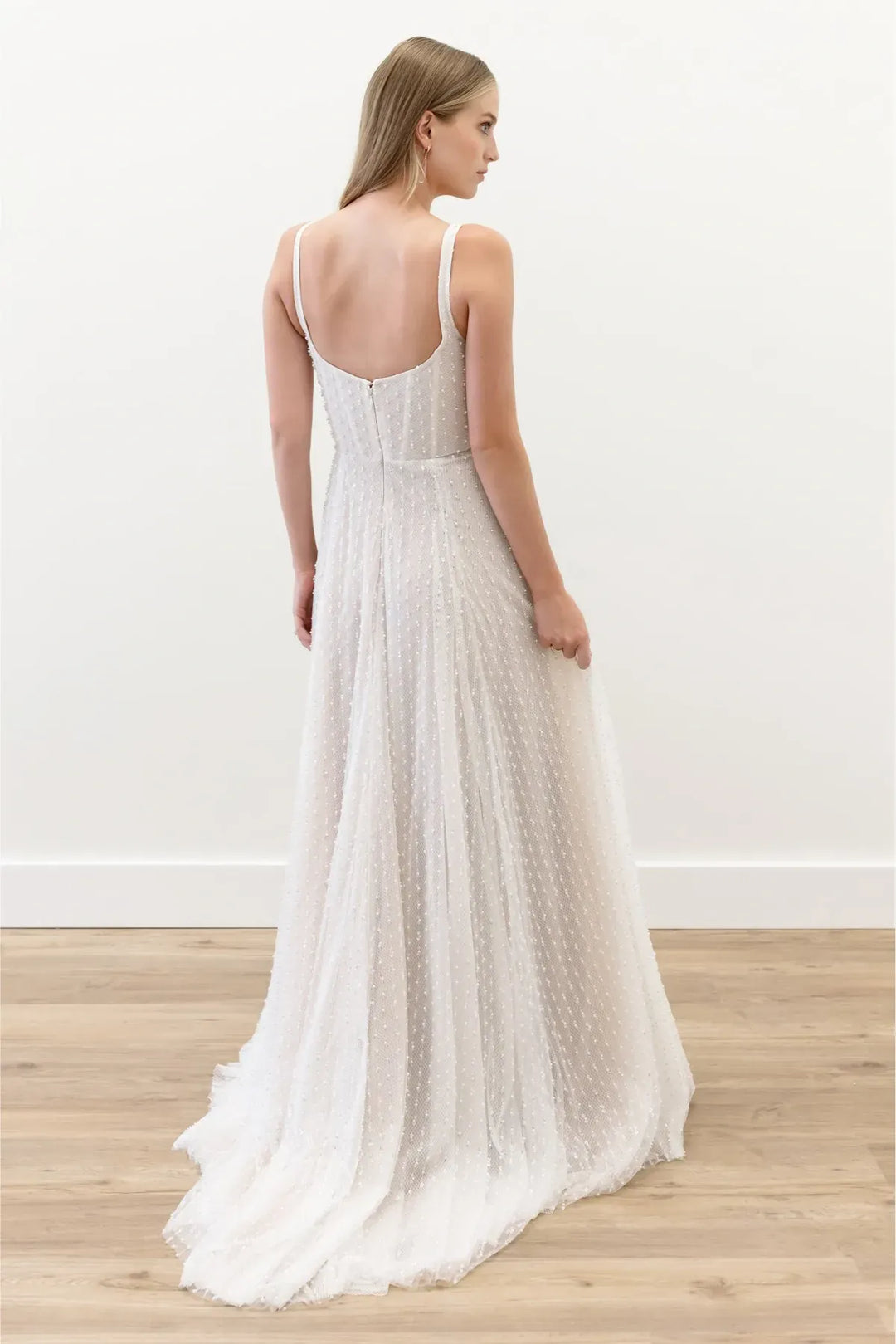 A woman wearing a sleeveless white maxi dress with thin straps, a fitted bodice, and a flowing textured skirt, shown from the back in a bright indoor room with a light wood floor.