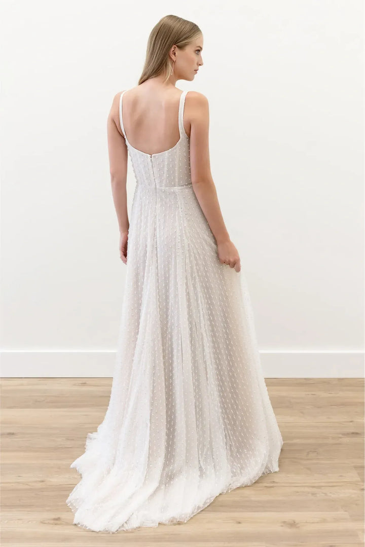 A woman wearing a sleeveless white maxi dress with thin straps, a fitted bodice, and a flowing textured skirt, shown from the back in a bright indoor room with a light wood floor.