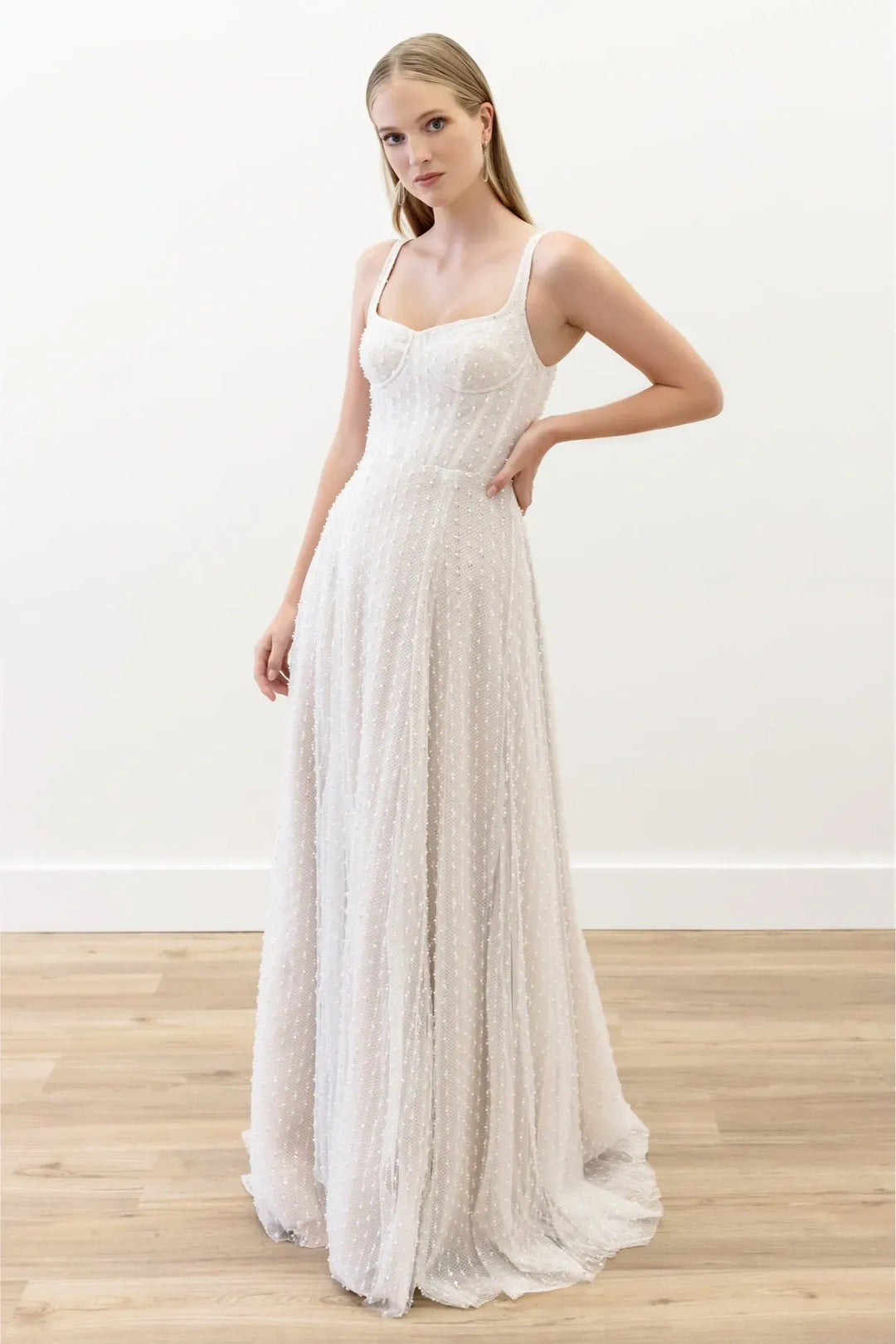 A woman modeling a sleeveless, floor-length white textured gown with a fitted bodice and flowing skirt, standing in a bright indoor room with a white wall and light wood floor.