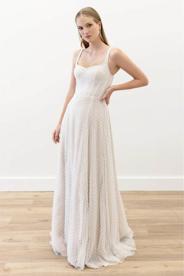 A woman modeling a sleeveless, floor-length white textured gown with a fitted bodice and flowing skirt, standing in a bright indoor room with a white wall and light wood floor.