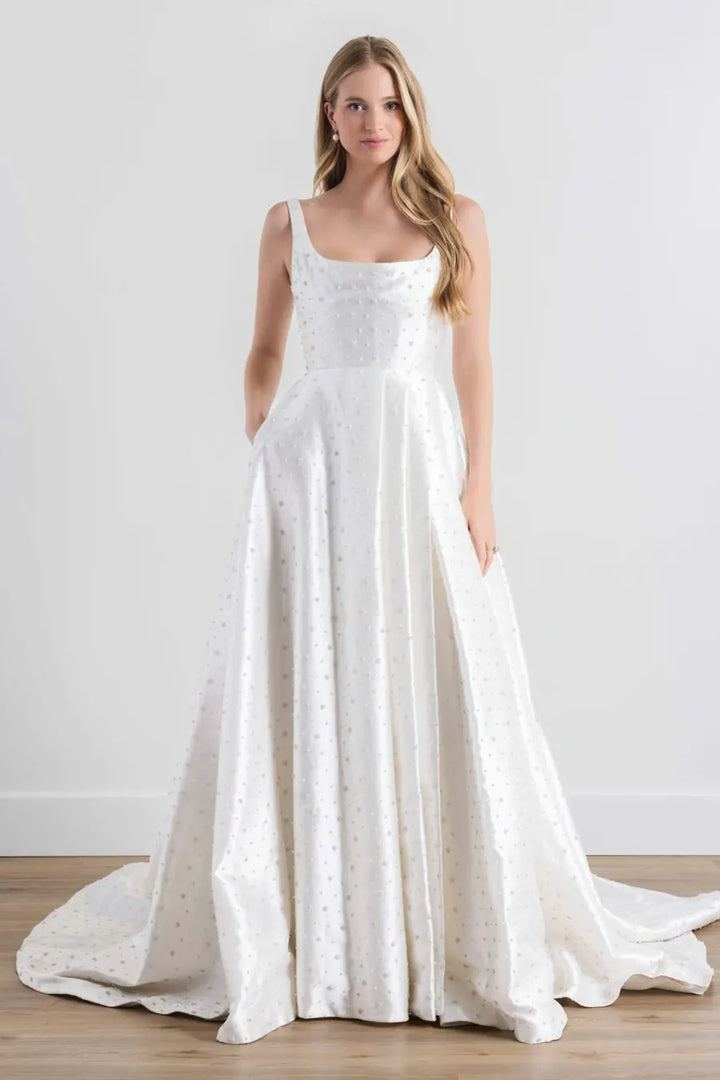 A woman modeling a white sleeveless A-line wedding gown with a fitted bodice, sheer embroidered overlay, side pockets, and a slight train on a light studio background.