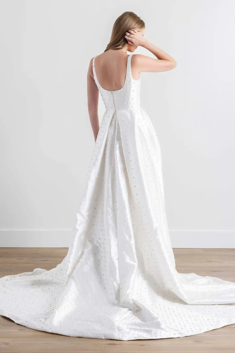 A woman wearing a white sleeveless wedding dress with a structured back and a long train, photographed in a bright indoor room with a light wall and wood floor.