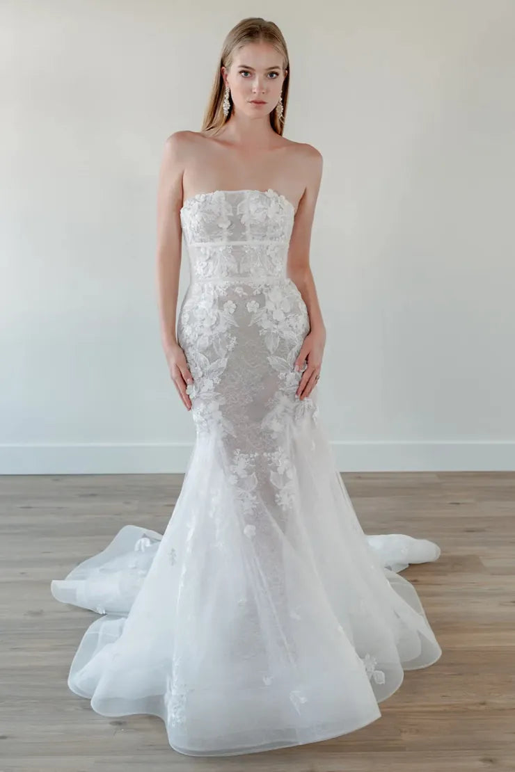 A strapless white lace mermaid-style wedding gown with a fitted bodice, sheer lace overlay, and a full layered tulle skirt with a train, shown on a model in a bright indoor room with light wood flooring.