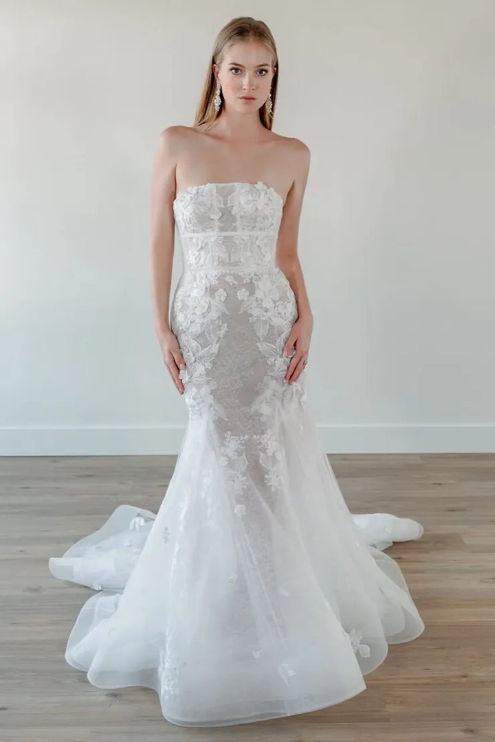 A strapless white lace mermaid-style wedding gown with a fitted bodice, sheer lace overlay, and a full layered tulle skirt with a train, shown on a model in a bright indoor room with light wood flooring.