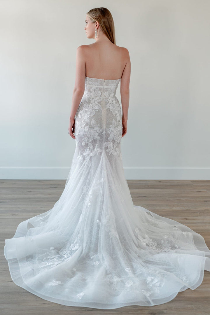 A woman modeling a strapless white wedding gown from the back, showing a fitted lace mermaid silhouette with a long sheer tulle train on a light wood floor against a plain off-white wall.