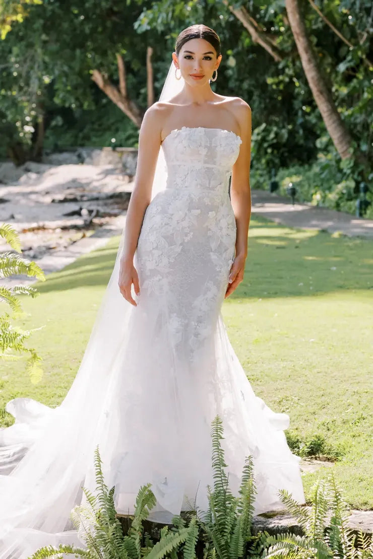 A woman wearing a strapless white lace mermaid-style wedding gown with a long sheer train, standing outdoors on a grassy lawn with trees and plants in the background.