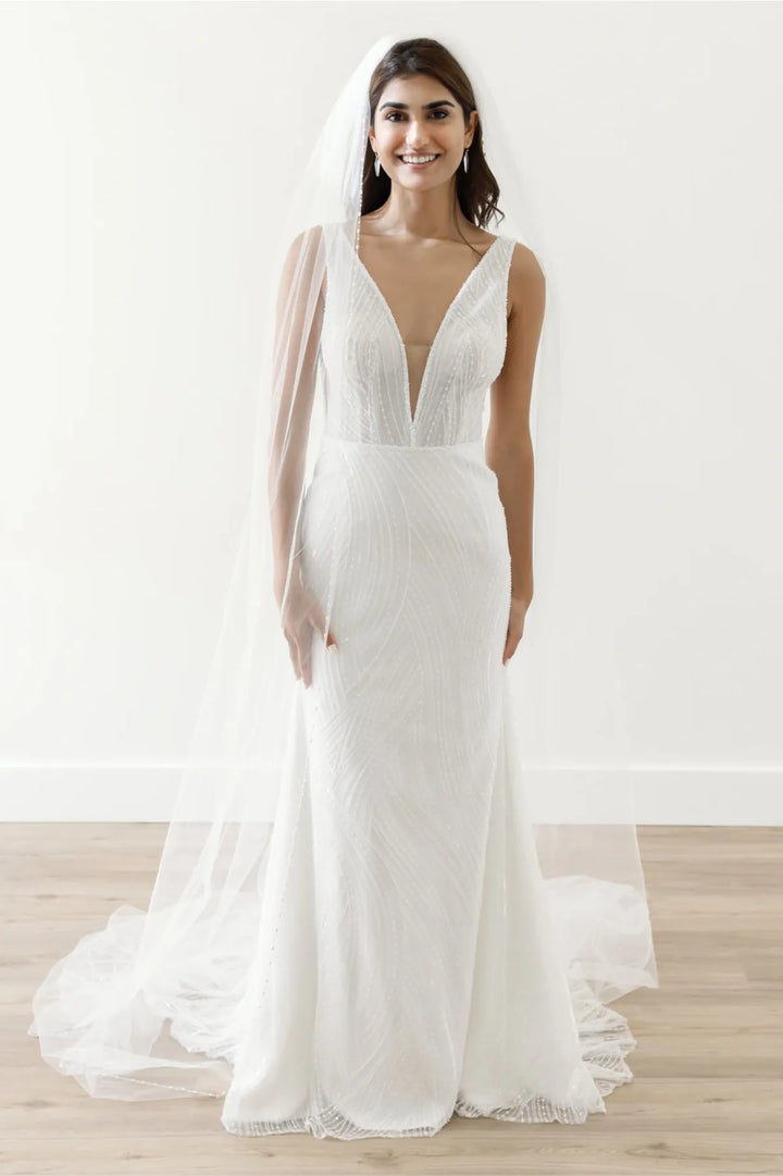 A woman modeling a white wedding gown with a deep V-neckline, fitted silhouette, sheer draped sleeve, and long sheer veil, standing on a light wood floor against a white wall.
