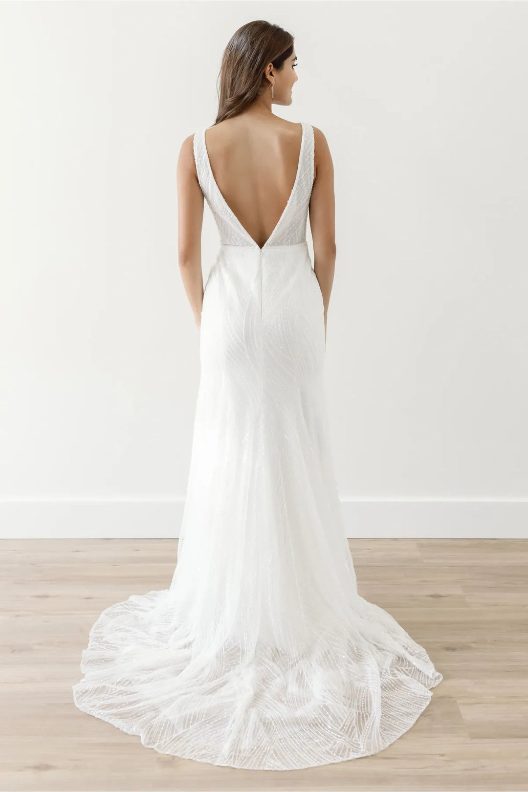 A woman wearing a white sleeveless wedding dress with a deep V back and a slight train, photographed from the back in a bright indoor studio with a light wall and wood floor.