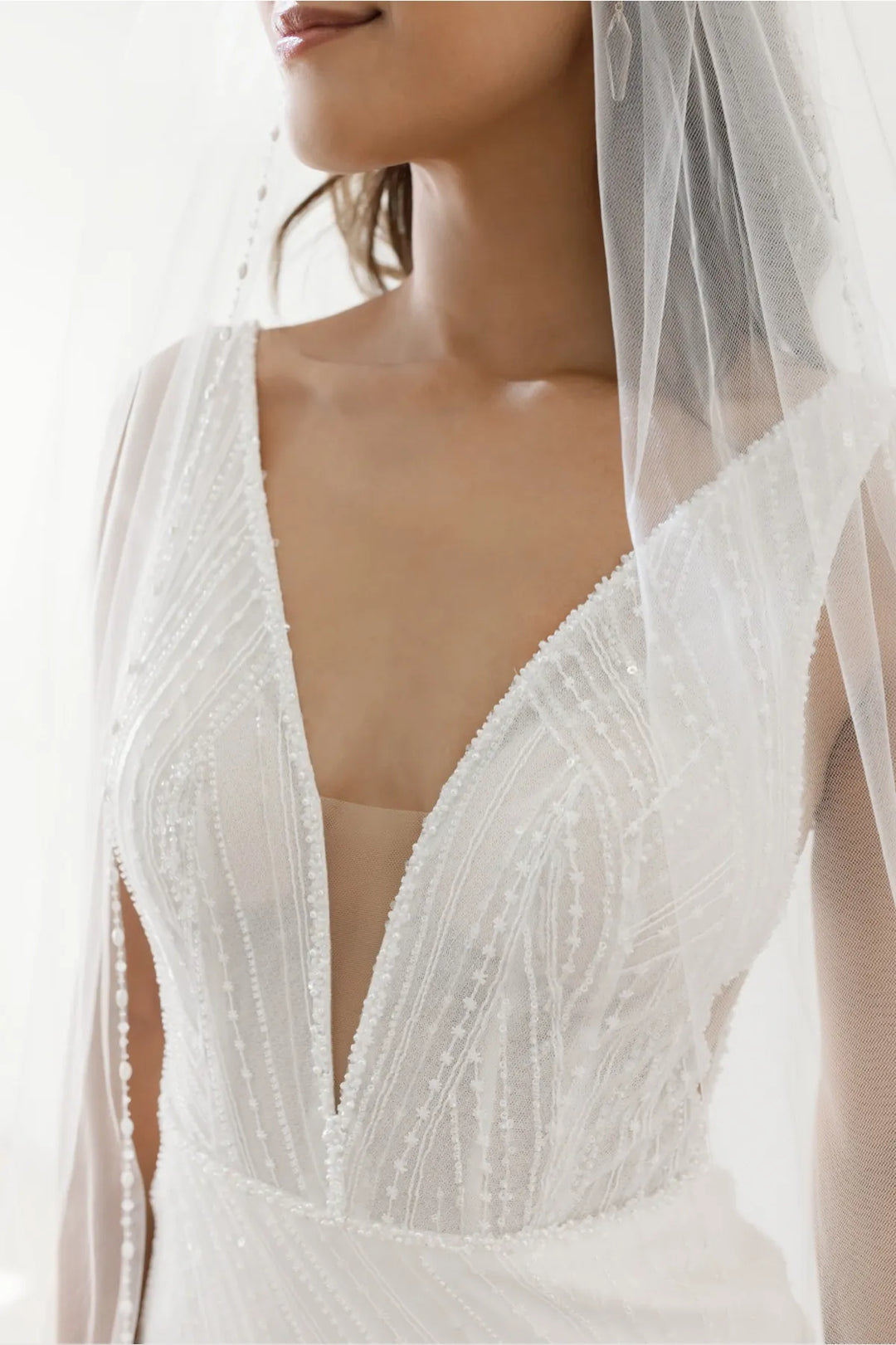 Close-up of a bride wearing a white beaded wedding dress with a deep V-neckline and a sheer veil, shown against a bright white background.
