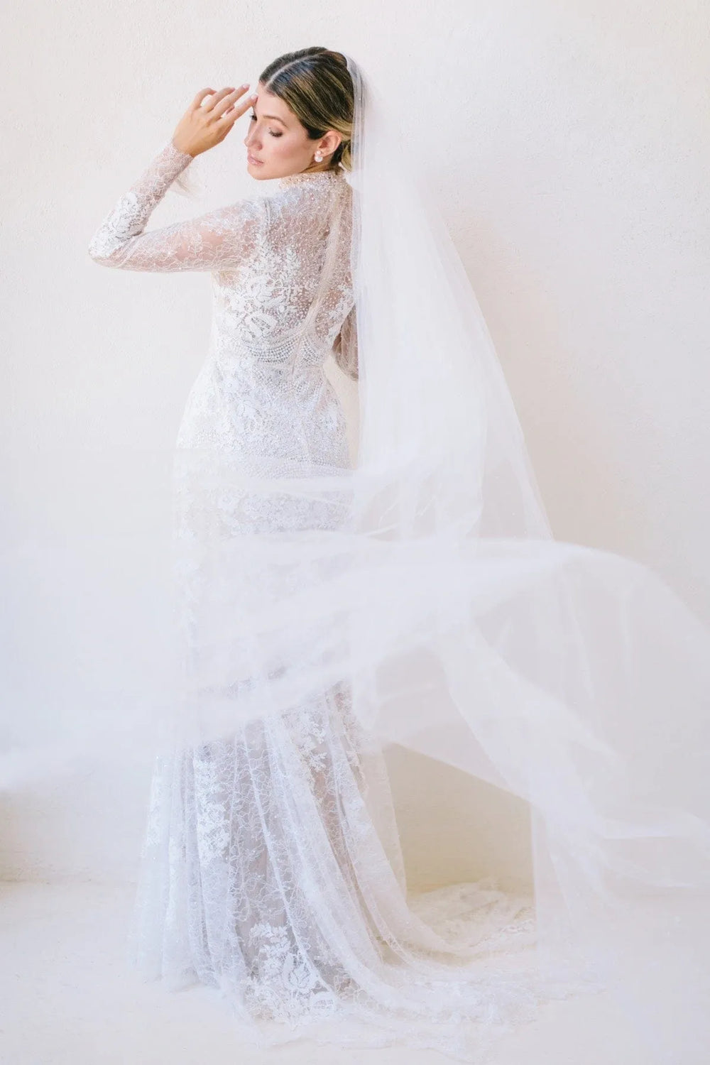 A bride wearing a long-sleeve lace mermaid-style wedding gown and a flowing sheer veil, posed against a soft cream studio background.