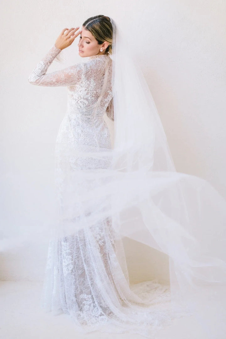 A bride wearing a long-sleeve lace mermaid-style wedding gown and a flowing sheer veil, posed against a soft cream studio background.