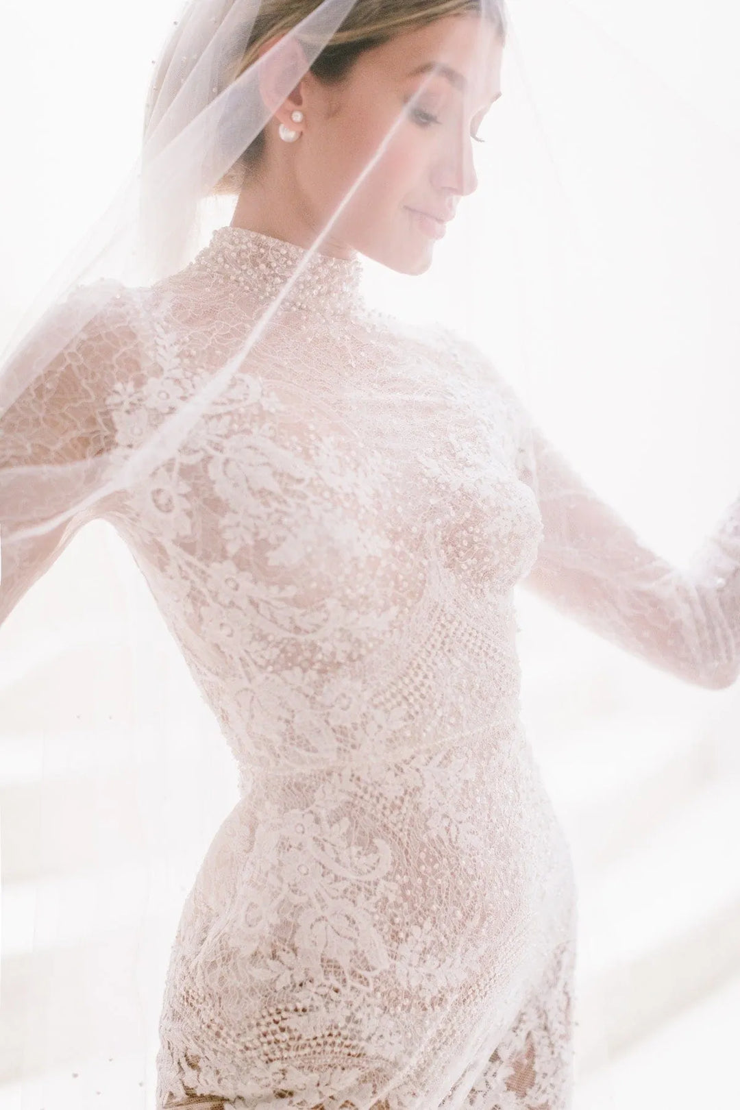 A woman wearing a sheer lace wedding dress with a high neckline and long sleeves, paired with a delicate veil, posed against a bright white background.