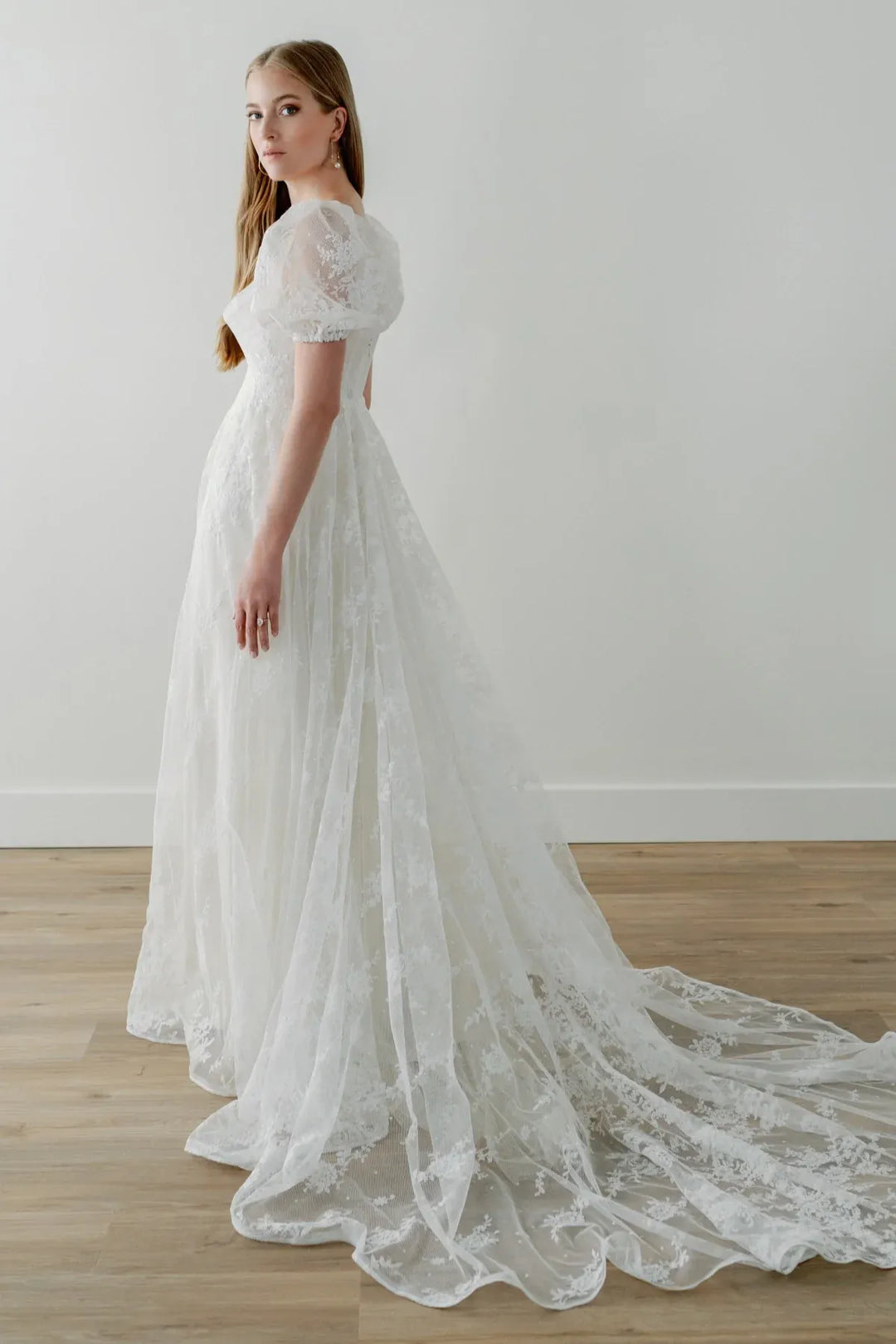 Woman wearing a white lace wedding dress with a long train on a wooden floor.