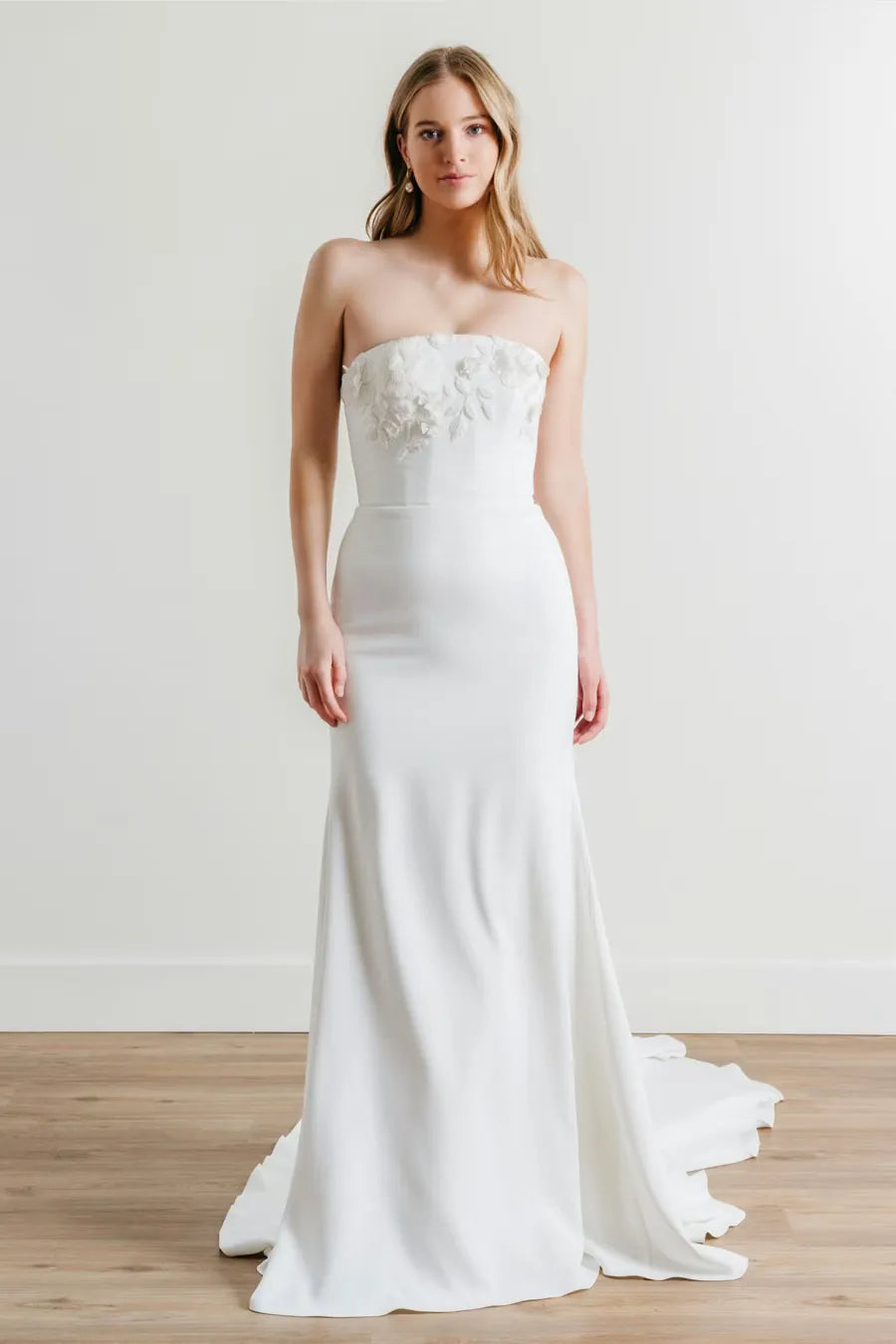 A woman modeling a strapless white wedding gown with floral applique across the bodice, standing in a bright indoor studio with a light wood floor and white wall.